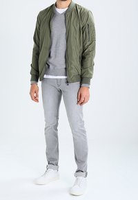 Veste bomber vert olive avec une poche zippée, pull gris porté en dessous, jean gris clair et baskets blanches. Tissu lisse, style décontracté.
