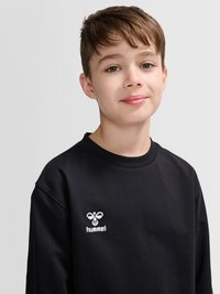 Jongen met bruin haar draagt een zwarte Hummel-trui en glimlacht lichtjes, tegen een effen lichtgrijze achtergrond.