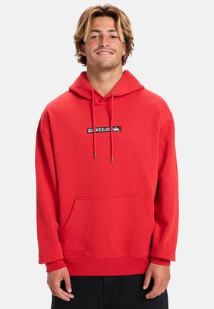 Roter Pullover-Hoodie aus weichem Material. Mit einer frontalen Känguru-Tasche und einem schwarzen Logo-Patch auf der Brust. Mit einer Kordelzug капюшон.