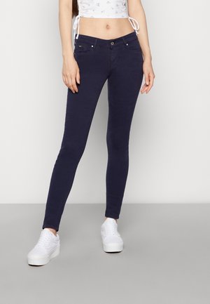 Donna che indossa jeans attillati blu navy, un top corto bianco a fiori e sneaker bianche con plateau, in piedi su uno sfondo neutro.