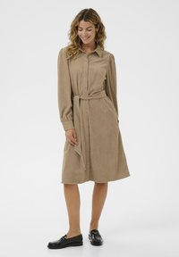 Beige corduroy shirt dress with long sleeves, button front, a tied belt at the waist, and a knee-length hem. Paired with black loafers.