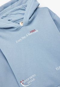 Sudadera de algodón azul claro con capucha ajustable y varios textos impresos en rojo y blanco, incluyendo "1985" y "ENTRA EN ESTILO".