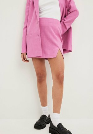 Vrouw draagt een roze blazer en rok met een voorscheur, witte sokken en zwarte chunky loafers tegen een lichte effen achtergrond.