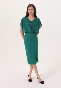 Robe verte avec une cape fluide et transparente, présentant un col orné de bijoux et une jupe ajustée jusqu'aux genoux avec une fente sur le côté. Escarpins noirs à bout pointu.