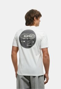 T-shirt in cotone bianco con una grande grafica circolare nera sulla schiena che rappresenta un globo e il testo "GUESS." Vestibilità casual, maniche corte.