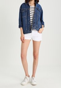 Tummansininen farkkutakki, napitettu, jossa on kaksi rintataskua. Raitapaita alla, valkoiset shortsit ja valkoiset lenkkitossut täydentävät asua.