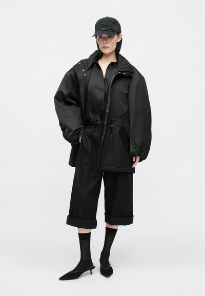 Crna oversized jakna s kapuljačom, uparena s crnom košuljom na dugmad i skraćenim hlačama. Dodatna oprema uključuje kapu i cipele s oštrim vrhom.