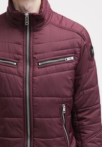 Vínová prošívaná bunda s vysokým límcem, která má dvě zipové náprsní kapsy a jednou zipovou kapsu po straně. Hladká látka s prošívanou texturou.