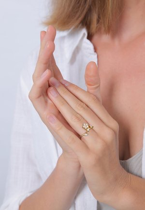 Goldring mit kleinem floralem Design und klaren Steinakzenten. Person, die die Hände zusammenhält und die Einzelheiten und Merkmale des Rings zeigt.