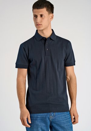 Mężczyzna w czarnej koszulce polo z krótkim rękawem w pionowe paski i niebieskich dżinsach, stojący na tle jasnego, jednolitego tła.