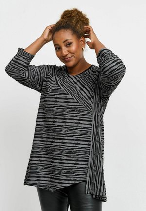 Femme aux cheveux bouclés portant une tunique à manches longues rayée gris et noir, ajustant ses cheveux, debout devant un fond clair uni.