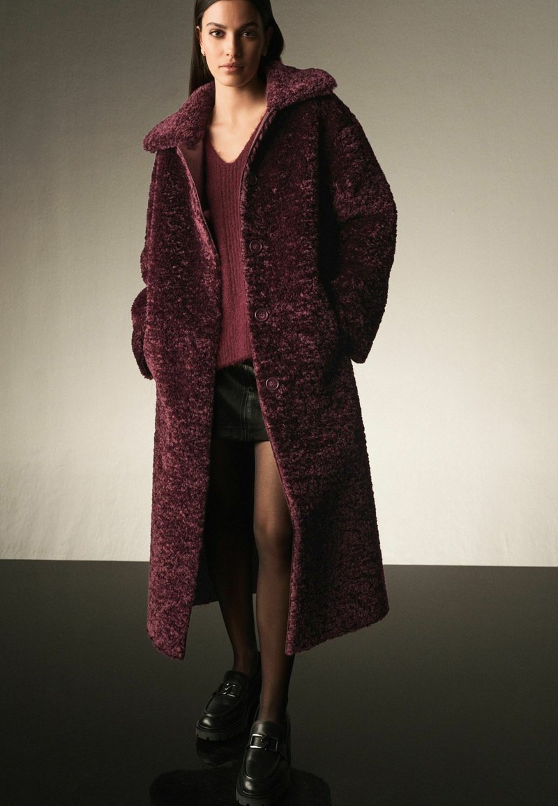 Lång, mörk bourgogne faux fur-kappa med bred krage, utrustad med knappstängningar och sidofickor, i kombination med en texturerad tröja och mörk kjol.