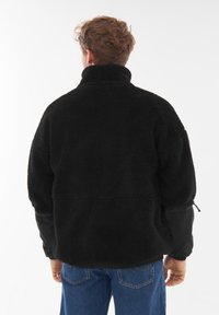 Giacca in pile nera con colletto alto, superficie testurizzata e polsini con zip, abbinata a jeans blu. La vista posteriore mette in evidenza il materiale e la vestibilità.