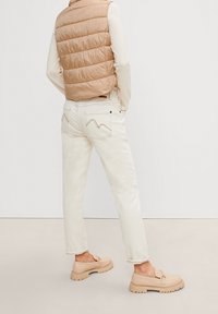 Béžová prošívaná vesta, krémové džíny se zickzackovým stehem na kapsách a světle hnědé boty s robustní podrážkou.