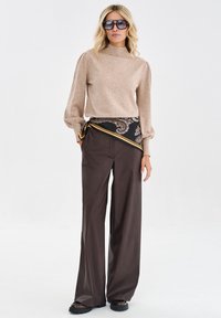 Beżowy sweter robiony na drutach z wysokim kołnierzem, w połączeniu z brązowymi spodniami o szerokich nogawkach. Brązowy szalik w wzory związany w talii. Duże czarne okulary przeciwsłoneczne.