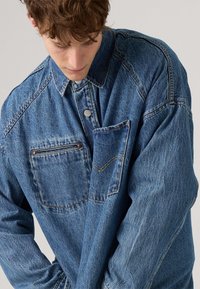 Blå denimskjorte med knap-up krave, oversized pasform, enkelt brystlomme og kontrasterende gul sømdetalj. Texturert stoff.