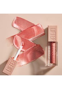 Le gloss Lifter de Maybelline dans un tube transparent, avec un applicateur blanc. Le gloss a une teinte rose brillante avec des échantillons lisses et brillants à proximité.