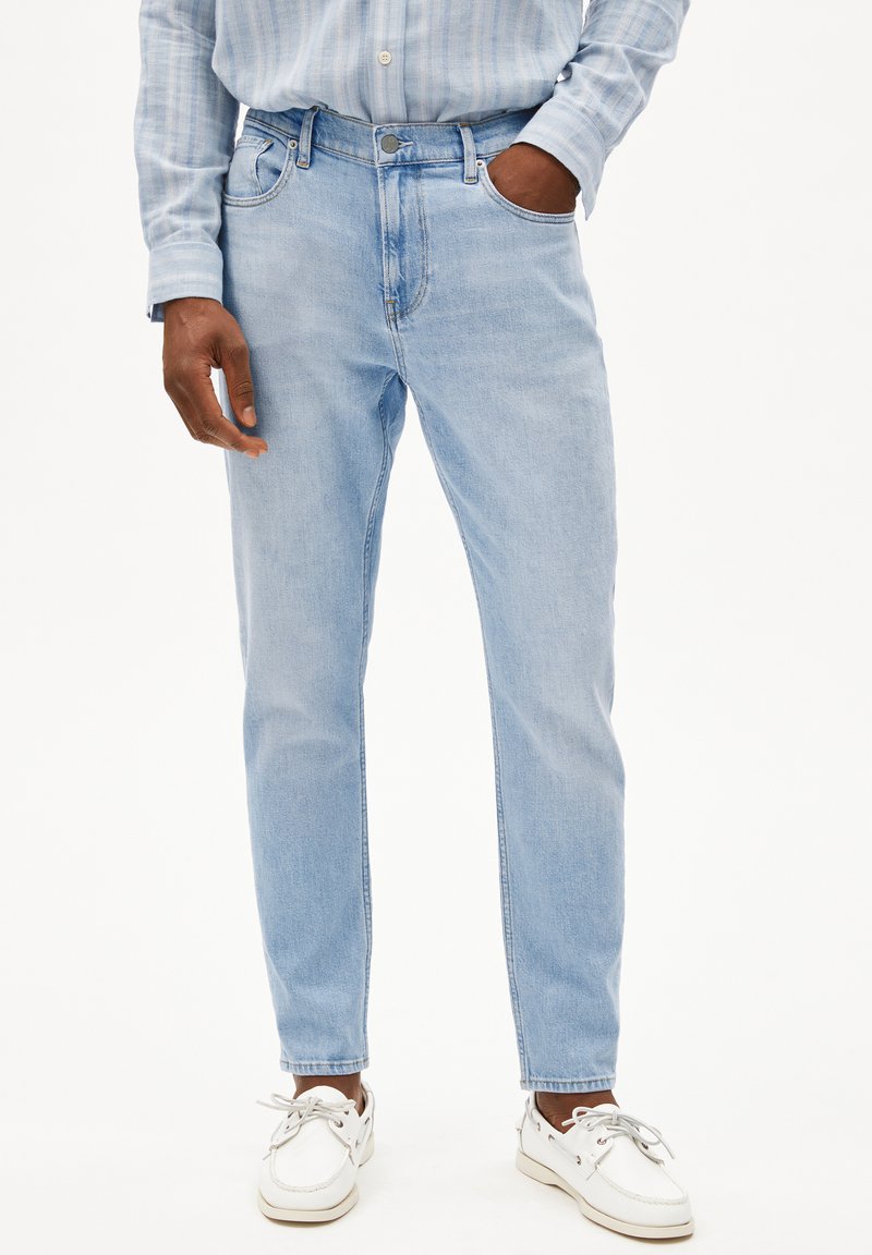 Jean en denim bleu clair avec une coupe droite, un design à cinq poches et une fermeture à bouton en métal, portés avec des chaussures bateau blanches.