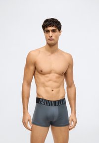Mies, joka ei ole paidassa ja jolla on lyhyet kiharat hiukset, pukeutuneena harmaisiin Calvin Klein -bokseritaskuille, seisoo yksinkertaista vaaleaa taustaa vasten.