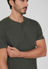 Ciemnozielona koszulka z krótkim rękawem, wykonana z miękkiego materiału, z okrągłym dekoltem i kieszonką na piersi. Gładka struktura i swobodny krój.