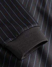 Tejido negro con rayas azul; cuenta con un puño acanalado para textura. El diseño liso y entallado acentúa la apariencia estructurada.