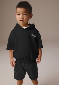Black oversized hoodie with short sleeves and ribbed hem, featuring a small white graphic on the chest, paired with matching black shorts.