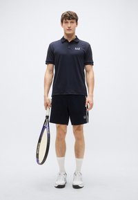 Temno modra polo majica z logotipom, združena s črnimi kratkimi hlačami z logotipom. Bele športne nogavice in čevlji. Drži teniški lopar.