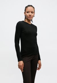 Top preto de manga comprida justo, feito de tecido liso, com decote redondo. Combinado com calças castanho-escuras justas.