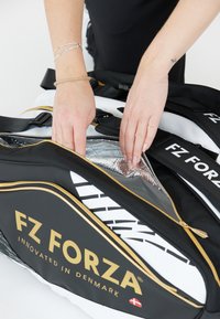 Svart och vit sportväska med gyllene detaljer; dragkedjan är öppen och avslöjar ett silverfärgat, isolerat fack. Logotypen "FZ FORZA" syns på sidan.