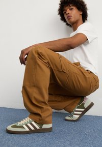 Ruske cargo hlače s više džepova, uparene s zeleno-sivim Adidas tenisicama s bijelim trakama. Jednostavna bijela majica.