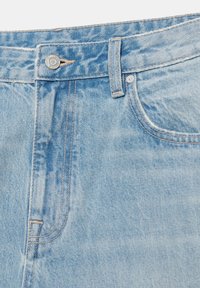 Jean en denim bleu clair en gros plan montrant le bouton, la passants de ceinture, la poche avant et les coutures détaillées.