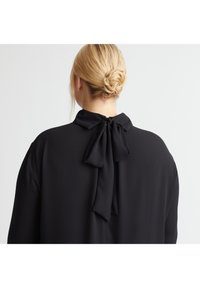 Schwarze Bluse mit einer Schleifen-Detail am Rücken, aus weichem Stoff, entspannter Passform und klassischem Kragen.