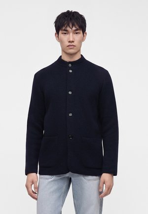 Junger Mann steht, trägt eine dunkel geknöpfte Jacke mit Fronttaschen und hellblaue Jeans vor einem einfarbigen weißen Hintergrund.
