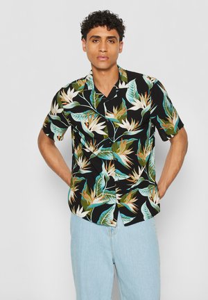 Mann steht mit Händen in den Taschen, trägt ein schwarzes kurzärmeliges Hemd mit Knöpfen und großem tropischem Blatt- und Blumenmuster sowie hellblaue Jeans.