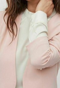 Ljusrosa blazer över en off-white blus. Blazern har en slät textur, skräddarsydd passform och slagna kragar. Blusen har en rundad krage.