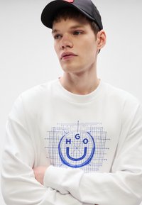 Λευκό φούτερ με μπλε γραφικό σχέδιο που απεικονίζει ένα χαμογελαστό πρόσωπο και τα γράμματα "HGO." Το μοντέλο φοράει μαύρο καπέλο, με σταυρωμένα χέρια. Ομαλή υφή.