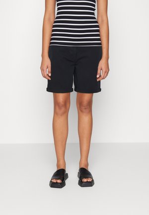 Personne portant un short noir à ourlet roulé, des sandales noires à bout ouvert, et un haut sans manches rayé noir et blanc sur un fond uni.