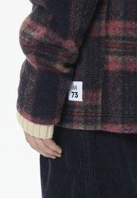 Sinine ja burgundi tooni ruuduline villane jakk, millel on silt suurusega 'M 73' ja kreemjat värvi kudumisega mansetid. All paistab vaibakanga kangas.