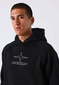Jonge man met kort, donker haar, die een zwarte hoodie draagt met een stippenster symbool en de tekst "PXLD" op de borst, met twee ringoorbellen.