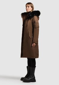 Abrigo largo marrón con capucha de piel sintética, que incluye una cremallera y bolsillos. Llevado sobre leggings negros y botas gruesas negras con cordones.