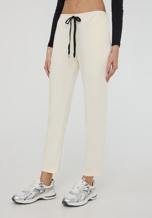 Pantaloni della tuta color crema realizzati in un tessuto morbido, con un cordino nero. Abbinati a scarpe da ginnastica argentate con dettagli a contrasto.