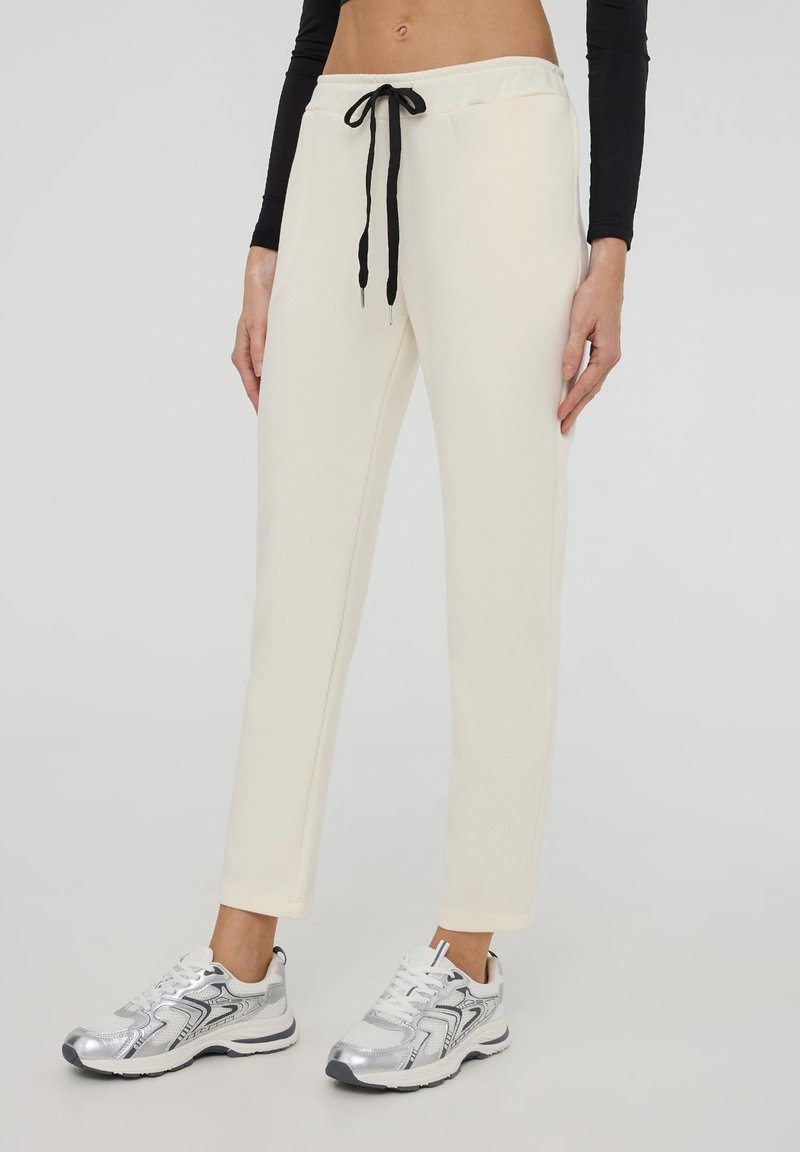 Pantaloni della tuta color crema realizzati in un tessuto morbido, con un cordino nero. Abbinati a scarpe da ginnastica argentate con dettagli a contrasto.