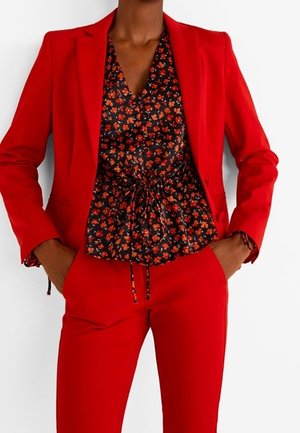 Personne portant un costume rouge vif avec un blazer et un pantalon sur mesure, associé à un chemisier noir à fleurs noué à la taille.