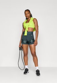 Neon zielony sportowy top związany w talii, ciemnoszare dopasowane szorty z napisem "NIKE PRO" na pasku, czarne sneakersy, trzymając skakankę.