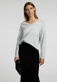 Lys grå striktrøje med V-hals og afslappet pasform, parret med en sort blyantskørt. Modellen holder en sort clutch i hånden.