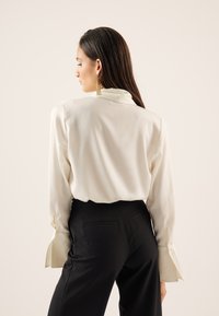 Bijela satenska bluza s dugim rukavima, s visokim ovratnikom i proširenim manžetama, u kombinaciji s crnim krojenim hlačama. Glatka tekstura.