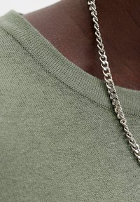 Pull tricoté vert olive à la texture douce, avec un col rond et un collier chaîne argentée visible à côté.