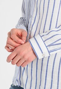 Camicia bianca di lino con righe verticali blu, dotata di polsini con bottone e colletto classico. Tessuto leggero e traspirante.