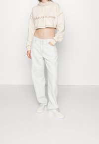 Felpa beige cropped con testo "Calvin Klein Jeans", abbinata a jeans a gamba larga blu chiaro e sneakers bianche. Texture del tessuto senza soluzione di continuità.