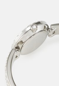 Silver stainless steel watch featuring a round case, textured bezel, and a crystal accent on the crown. Smooth metal strap with a clasp.